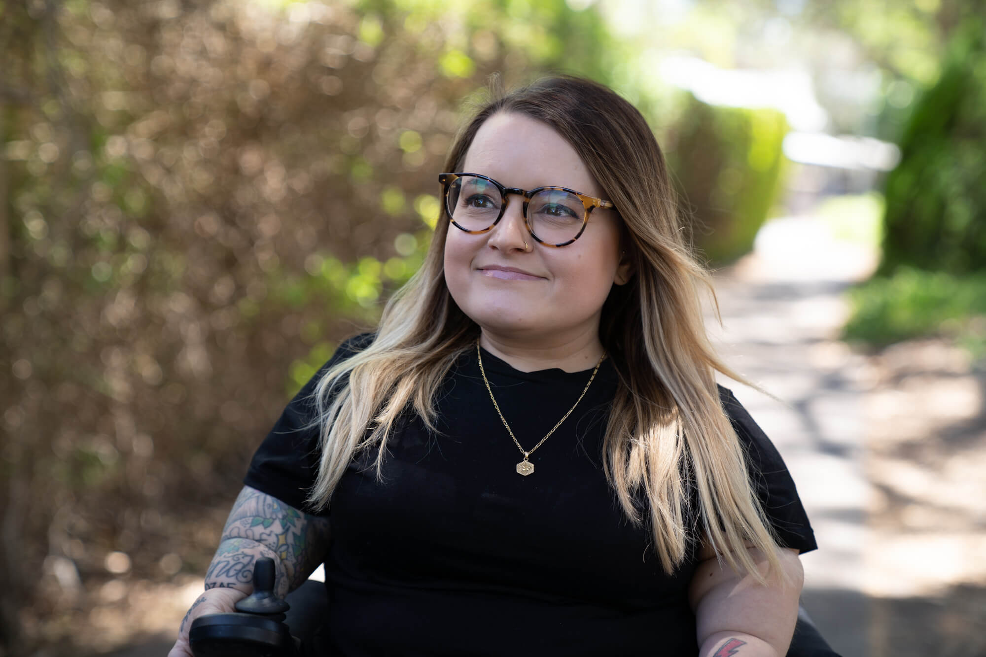 Belle outdoors Photo of Belle in her motorised wheelchair outdoors. She wears a short sleeved black shirt which shows her tattooed sleeve. She has tortoiseshell framed glasses and dark brown hair with blonde tips down to her shoulders, and is smiling.