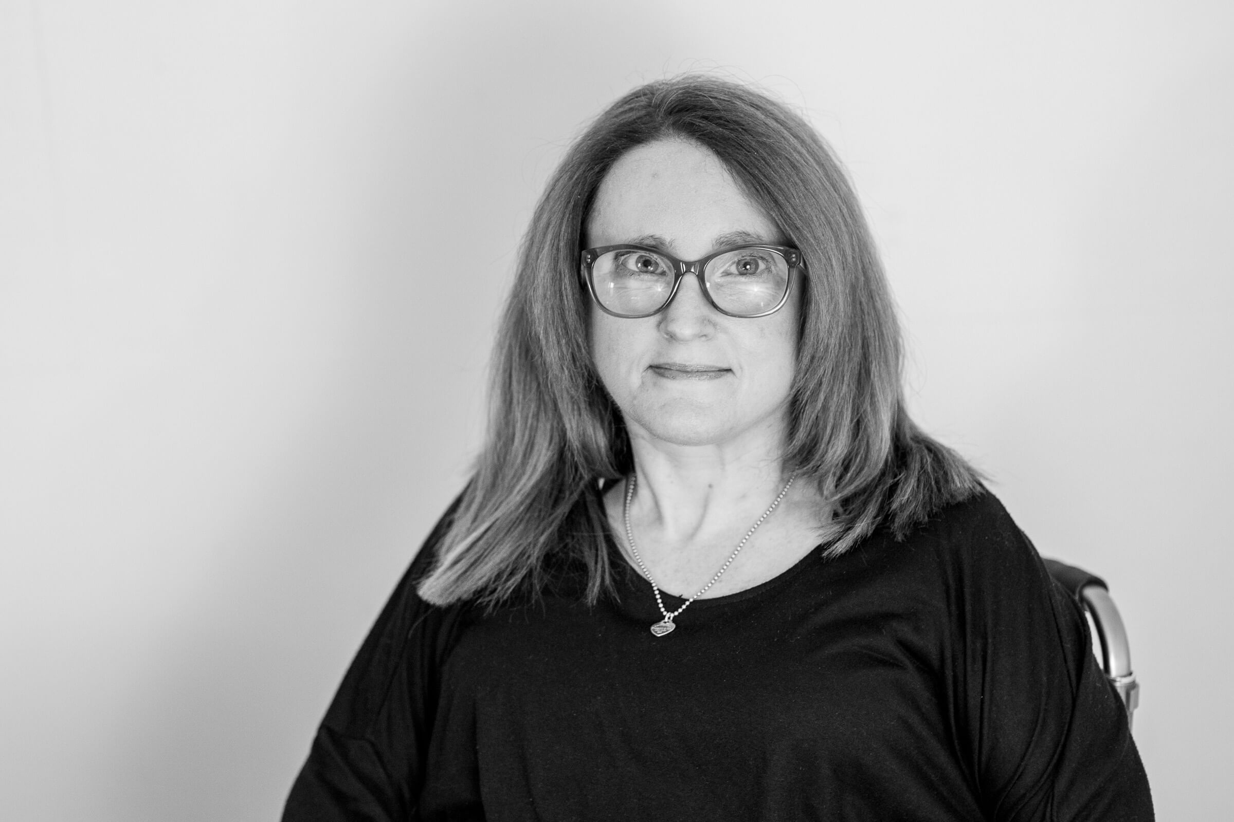 Black and white photo of Ainslee A black and white photo of Ainslee looking at the camera. She has shoulder length straight hair framing her face, wears glasses and a necklace, and a dark coloured top.