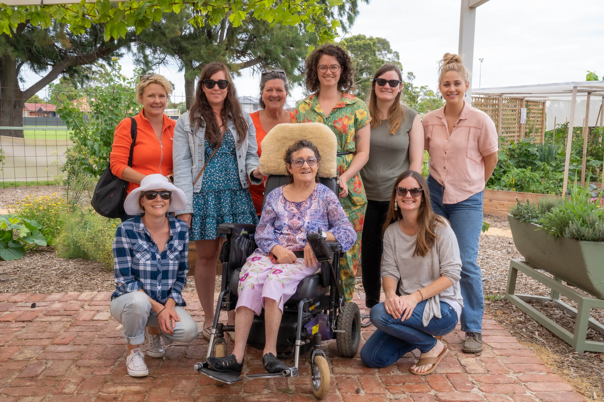 Photo is a group shot of 9 women all smiling at the camera. Sally is in the middle in her wheelchair, Katie is couched to her left and Rebecca to her right. Behind them, from left to right, stand Renee, Jess, Colleen, Elizabeth, Nicole and Alex.