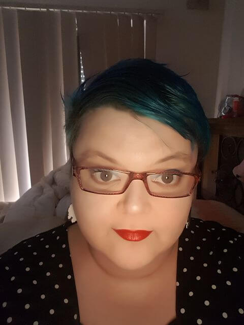 A selfie of Eva, who has short cropped turquoise hair, wears red lipstick and glasses with red frame.