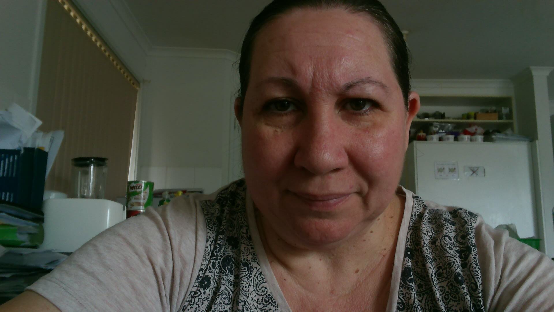 Selfie of Donna A selfie of Donna, with the background of a kitchen. She is a woman with dark hair tied back, wears a grey top with a black swirl pattern in the front, and is looking straight into the camera.