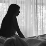 Photo of a sad woman sitting by the window in a bedroom.