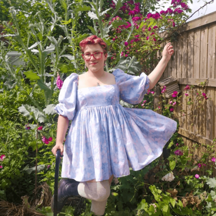 Zoë, a petite white person with short pink hair is stood in a garden filled with foxgloves, roses, giant ornamental thistles, sweet peas, and foliage. They wear a pastel blue empire dress with puffed sleeves and short flared skirt. They wear white stockings and lace up black boots, leaning on a black walking stick smiling.