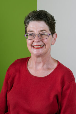 Photo of Annette smiling at something off-camera. Photo of Annette smiling at something off-camera. She is a white woman with glasses and short dark hair, wearing a red long sleeved shirt, standing against a split green and grey background.