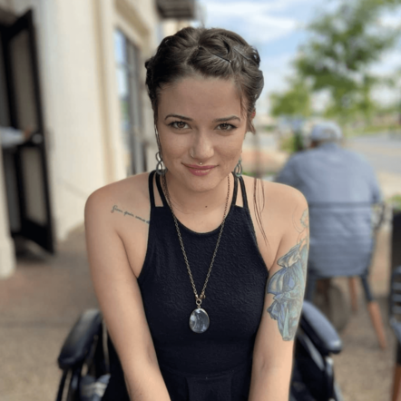 Photo of a white woman with dark hair in an updo smiling straight at the camera, while sitting in a black wheelchair. She has a chunky gem necklace and wears a sleeveless black top. Behind her is a blurred background of a building and someone seated in a chair outdoors, trees in the distance.