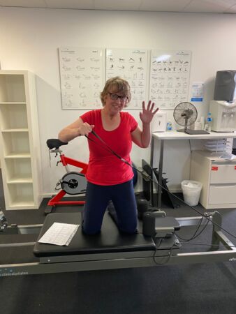 Jane having fun in her pilates class. She is kneeling down on a pilates reformer and waving to the camera.