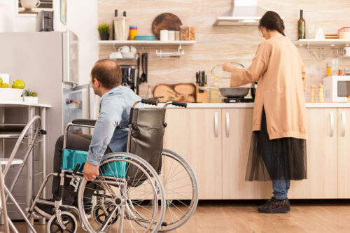 Photo of wheelchair user opening fridge. Photo shows man in wheelchair opening a fridge, while a woman cooks in the background.
