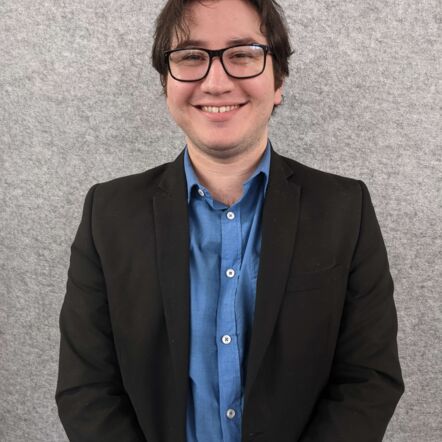 Photo of Dylan, a young man with dark hair and white skin, wearing glasses, a blue collared shirt and a black suit. He is standing against a grey wall and smiling at the camera.