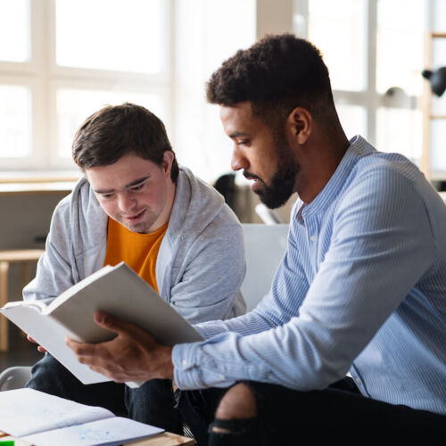 Photo shows a man with Downs syndrome and African man looking at an open book together.