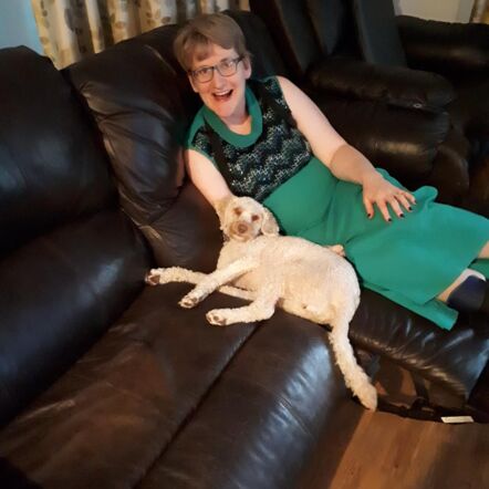Photo of Melissa on a black sofa with a pet dog. She is a woman with brown, short cropped hair wearing an emerald dress with black lace detail on the top. She is smiling at the camera.