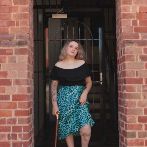 Photo of Lea standing in a doorway flanked by brick walls. She has long blonde hair, wears a black off shoulder top and blue leopard print knee length skirt. She holds a walking stick, and you can see the scars on her left leg.