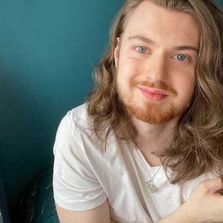 Photo of Callum smiling at the camera. He has white skin, blue eyes, light brown facial hair and long, light brown wavy hair that falls over his shoulders. He is wearing a white V-neck shirt and a pendant dangles from his neck. The background is a solid teal colour.