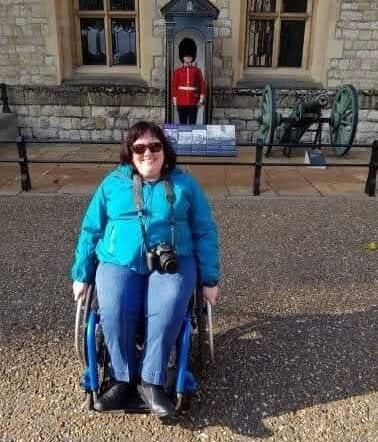 Photo of Jorja on her trip to the UK. She is wearing sunglasses, a bright blue long sleeved top and jeans. There is a camera around her neck,and she is in front of a display of the Queen's Guard.