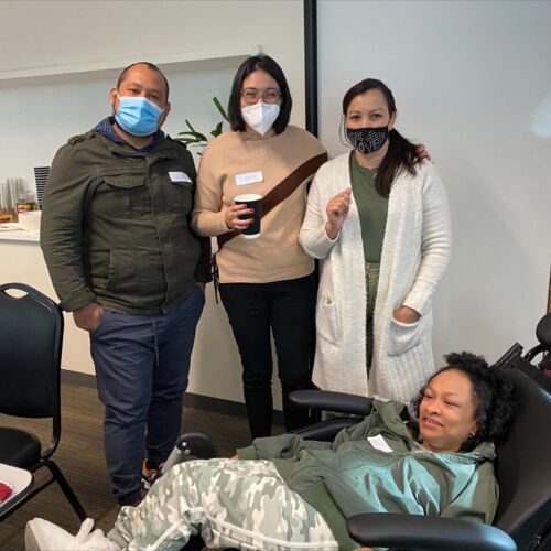Photo shows a group of 3 adults standing, a man and two women, their nose and mouths covered with masks, but their eyes lit up with smiles, and a child with dark curly hair, leaning back in a reclined black chair.