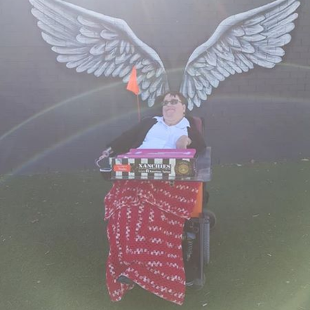Nadia sitting in a wheelchair with a red and white crochet blanket over her lap. She is dressed in a pale blue shirt with a collar with a dark cardigan over the shirt. She is holding a carton with her right hand, which is a hook. In the background is a painting of angel wings on a brick wall, so it looks like Nadia has wings.