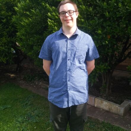 Photo of Joseph outdoors, smiling at the camera. He has short brown hair, black glasses and is wearing a collared blue shirt and cargo pants.