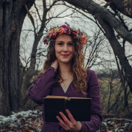 Photo of Rebekah outdoors in a barren forest, snow lightly covering the forest floor. She is a woman with white skin, a flower crown atop her head, long light brown curly hair cascading below her shoulders, and wears a long sleeved deep purple top. She holds an open book with one hand and the other hand touches the back of her neck.