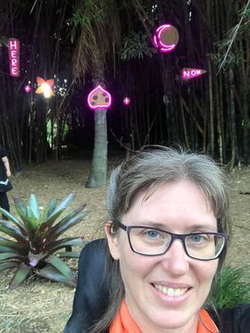 Selfie of Sarah, a woman with white skin, purple framed glasses and light brown hair, smiling at the camera. In the background is a bamboo grove and single palm tree, with neon lights of various shapes and words "Here" and "Now" on the trunks.