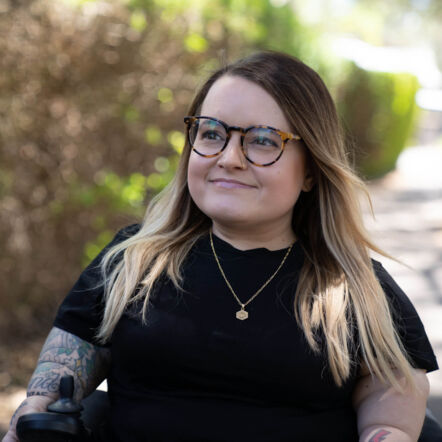 Photo of Belle in her motorised wheelchair outdoors. She wears a short sleeved black shirt which shows her tattooed sleeve on her right arm. She has tortoiseshell framed glasses and dark brown hair with blonde tips down to her shoulders, and is smiling at something in the distance.