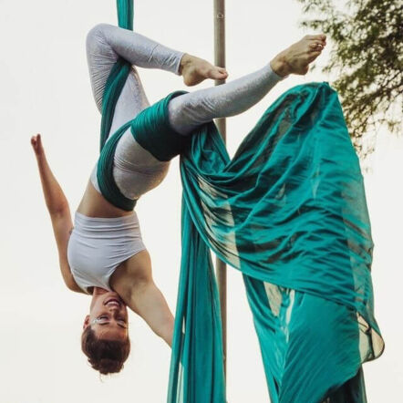 Photo of Sarah upside down in the air, held up by emerald green fabrics wrapped behind her knees and around her waist. She has her dark hair in an updo, sparkly silver glitter around her eyes and wears a sleeveless white crop top and silver leggings. She is smiling, both arms spread out, fingers pointed. One hand is smaller than the other. Her feet are pointed, and one knee is bent at an angle towards the calf of her other leg.