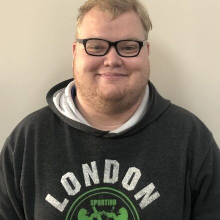 Photo of James smiling straight to the camera. He is a man with light blonde hair and stubble on white skin, wearing a black hoodie with the words "London Sporting", black glasses framing his eyes.