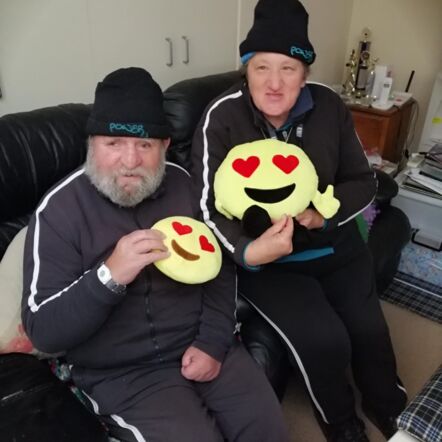 Mr Peter and Mrs Debbie W at home, sitting on the sofa. They are dressed in matching dark trackies and beanies that say "Power" in teal text. Both are looking at the camera and holding cushions shaped like the heart eyes emoji.