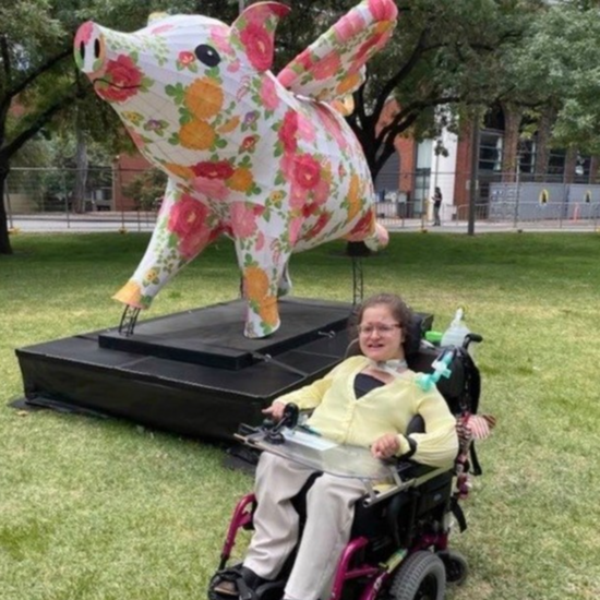 Photo of Alice outdoors in her wheelchair. Alice sits in her wheelchair outdoors on the grass. She wears a yellow top and glasses and is smiling. Behind her is a large art sculpture of a pig with wings, a pattern of red and orange flowers across its body.