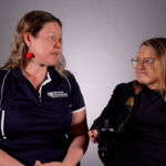 Two woman are in a conversation in front of a grey backdrop. One is using a wheelchair