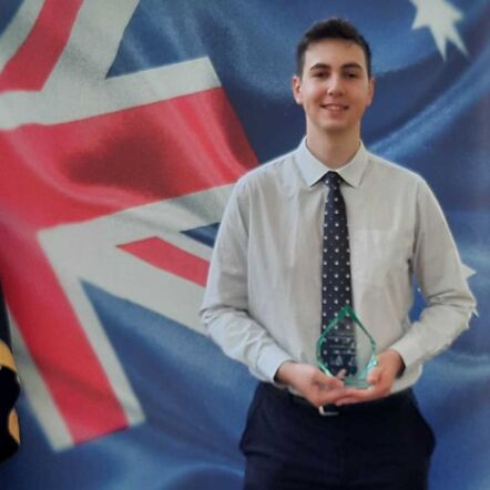 Photo of James holding his Award and standing in front of a flag of Australia. He has white skin, short dark hair and is dressed smartly in a collared shirt and tie.