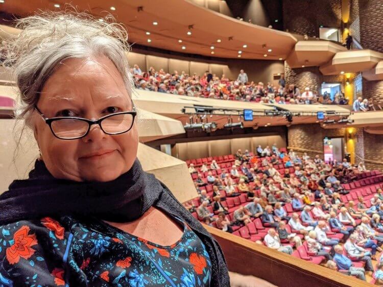 Julie in a crowded amphitheatre Photo of Julie indoors in a crowded amphitheatre. She has grey hair that's swept back, black framed glasses and wears a black top with a floral pattern and a black scarf around her neck.