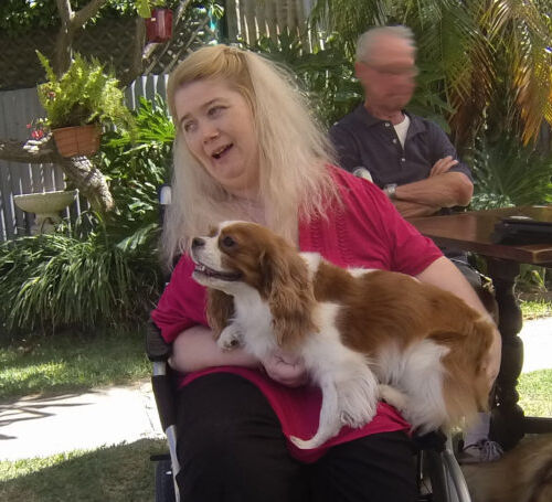 Photo of Ann Marie Smith in a garden. She has long light blonde hair down to her shoulders and is holding a dog in her lap, smiling.