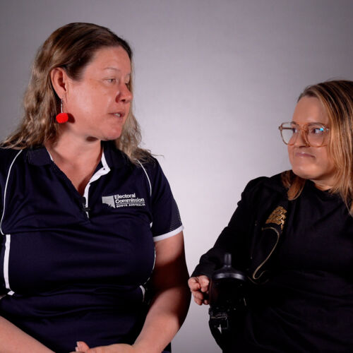 Two woman are in a conversation in front of a grey backdrop. One is using a wheelchair
