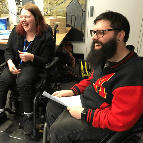 Photo shows 2 members of EYDN at a peer network meeting. On the left is Sarah laughing at a joke. She has pink streaks running through her light brown shoulder length hair and is dressed in black. To the right is Nick smiling. He has short dark hair and a beard, and is wearing a black jacket with red sleeves and a big letter "A" emblazoned  on the chest.