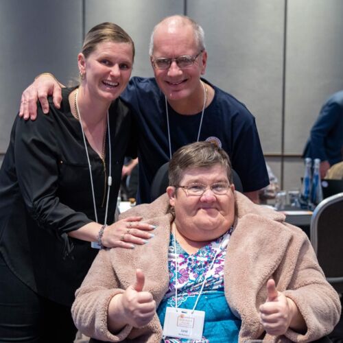 Photo shows 3 people at the OVSA conference. A man and woman are standing and smiling at the camera, with another woman seated at the table in front of them, giving a thumbs up to the camera.